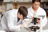 Scientist looking in a microscope while his colleague is taking notes