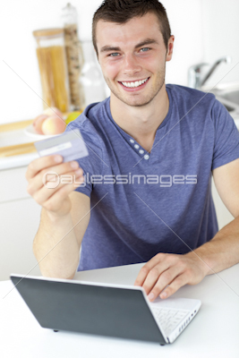 Charismatic young man holding a card using his laptop in the kitchen smiling at the camera