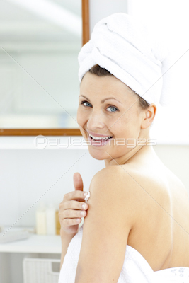 Delighted young woman putting cream on her body looking at the camera