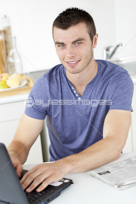 Handsome young man using his laptop looking at the camera in the kitchen