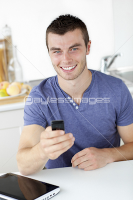 Lively young man sending a text smiling at the camera in the kitchen