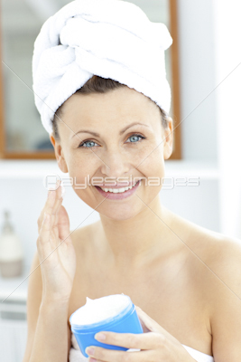 Pretty young woman putting moisturizer on her face looking at the camera