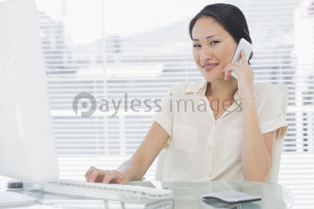 The female businessman is using the mobile phone and the computer at the desk.