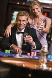 Couple+in+casino+at+roulette+table+holding+up+champagne+and+smiling+%28selective+focus%29