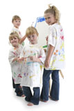 Four+girls+in+big+white+shirts+covered+in+paint+at+easel.+Shot+over+white+background.