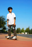 Teenage+boy+riding+a+skateboard+on+the+sidewalk+of+a+parking+lot+on+a+sunny+day+with+blue+sky+and+trees+in+the+background.+