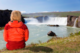 A+young+woman+sitting+looking+at+a+waterfall.+Shot+on+location+at+Godafoss+waterfall+in+Iceland.
