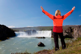 A+young+woman+with+her+arms+up+in+celebration+of+achievement+by+a+waterfall.+Shot+on+location+at+Godafoss+waterfall+in+Iceland.