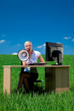 Business+concept+shot+of+a+beautiful+young+woman+sitting+at+a+desk+using+a+megaphone+in+a+green+field+with+a+bright+blue+sky.+Shot+on+location.