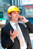 A+man+in+a+yellow+hard+hat+and+suit+on+an+industrial+or+constructin+site+talking+on+his+cell+phone