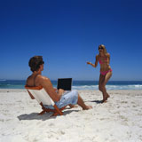 Young+man+using+a+laptop+on+the+beach+with+a+young+woman+laughing+in+front+of+him