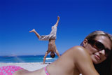 Close-up+of+a+young+woman+lying+on+the+beach+with+a+young+man+doing+a+handstand+in+the+background