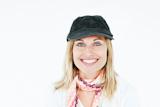 Bright blond woman with cap and scarf smiling at the camera against a white ط