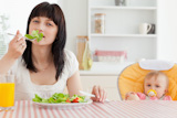 Attractive brunette woman eating a salad next to her baby while sitting