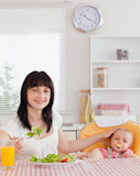 Pretty brunette woman eating a salad next to her baby while sitting