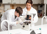 Scientist looking in a microscope while his coworker is taking notes